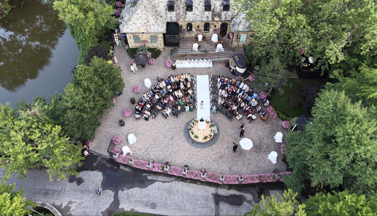 Wide aerial drone photograph of a wedding ceremony with guests seated in curved rows facing a stone patio and pond