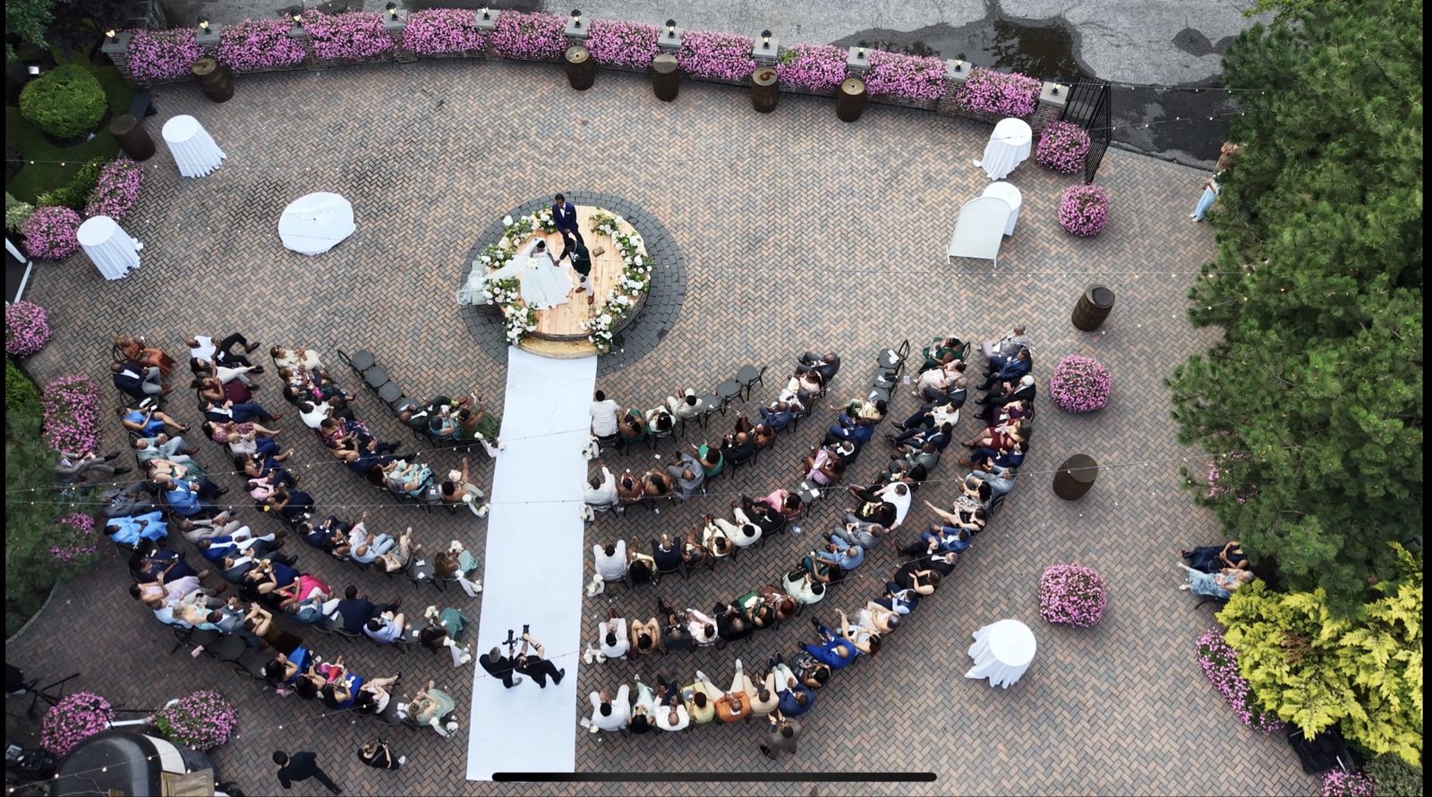 Aerial drone photograph of a couple at their wedding ceremony beneath a floral arch, surrounded by fan-shaped guest seating and string lights